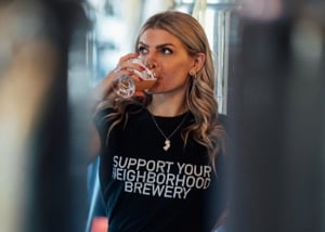 Maddie Bowen ’19 drinks from a glass while standing inside a brewery, wearing a shirt that reads “Support Your Neighborhood Brewery.”
